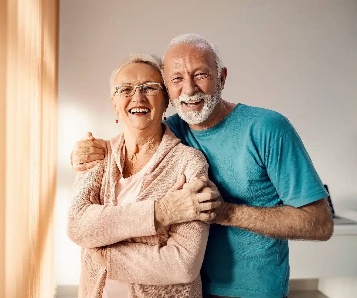 Senior couple hugging in a nursing home. A happy senior couple standing next to a window in a nursing home, hugging and smiling.